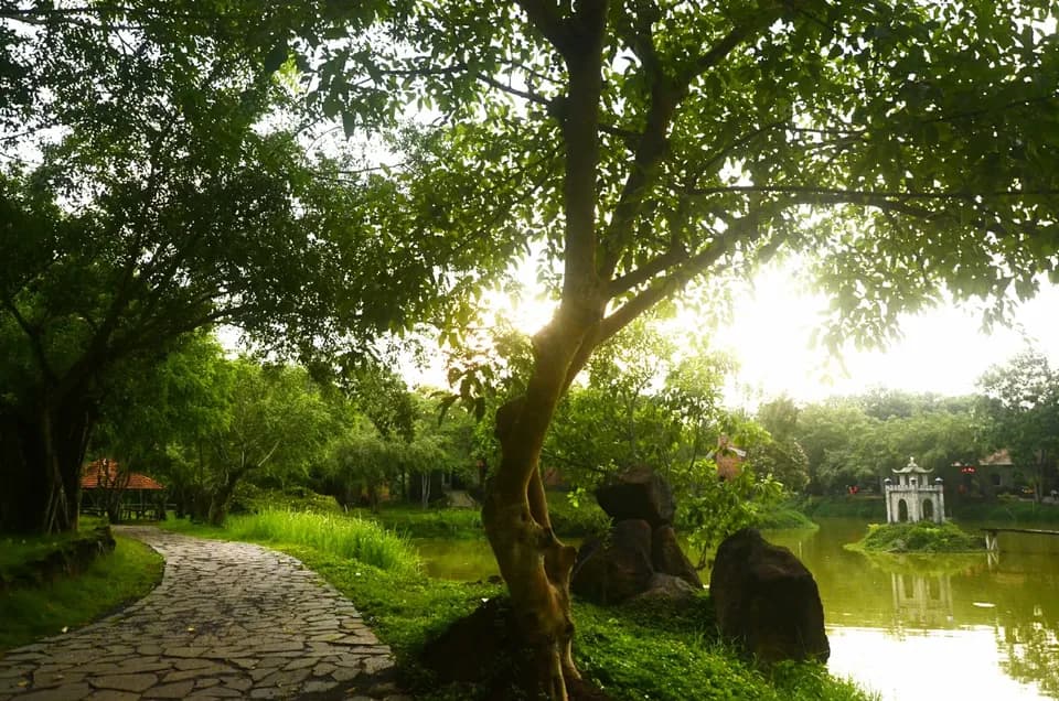 Lối đi lát đá uốn quanh hồ dưới tán cây, xa xa là ngôi miếu nhỏ bên mặt nước